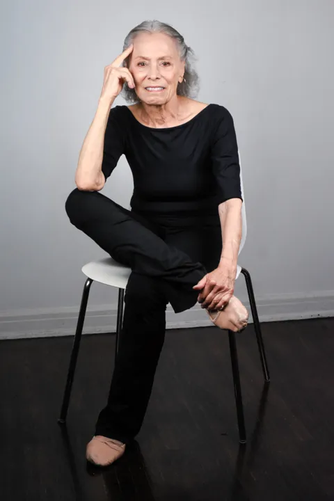 Michele Hart seated cross-legged on a stool in black dance clothes, hand resting at her temple